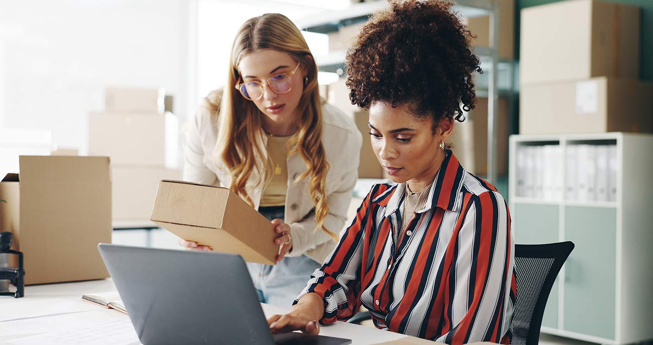 Two women looking at a laptop screen, surrounded by boxes. 